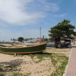 Imagem da Praia Central de Barra Velha, onde logo a frente há um barco, com natureza e praia ao fundo,